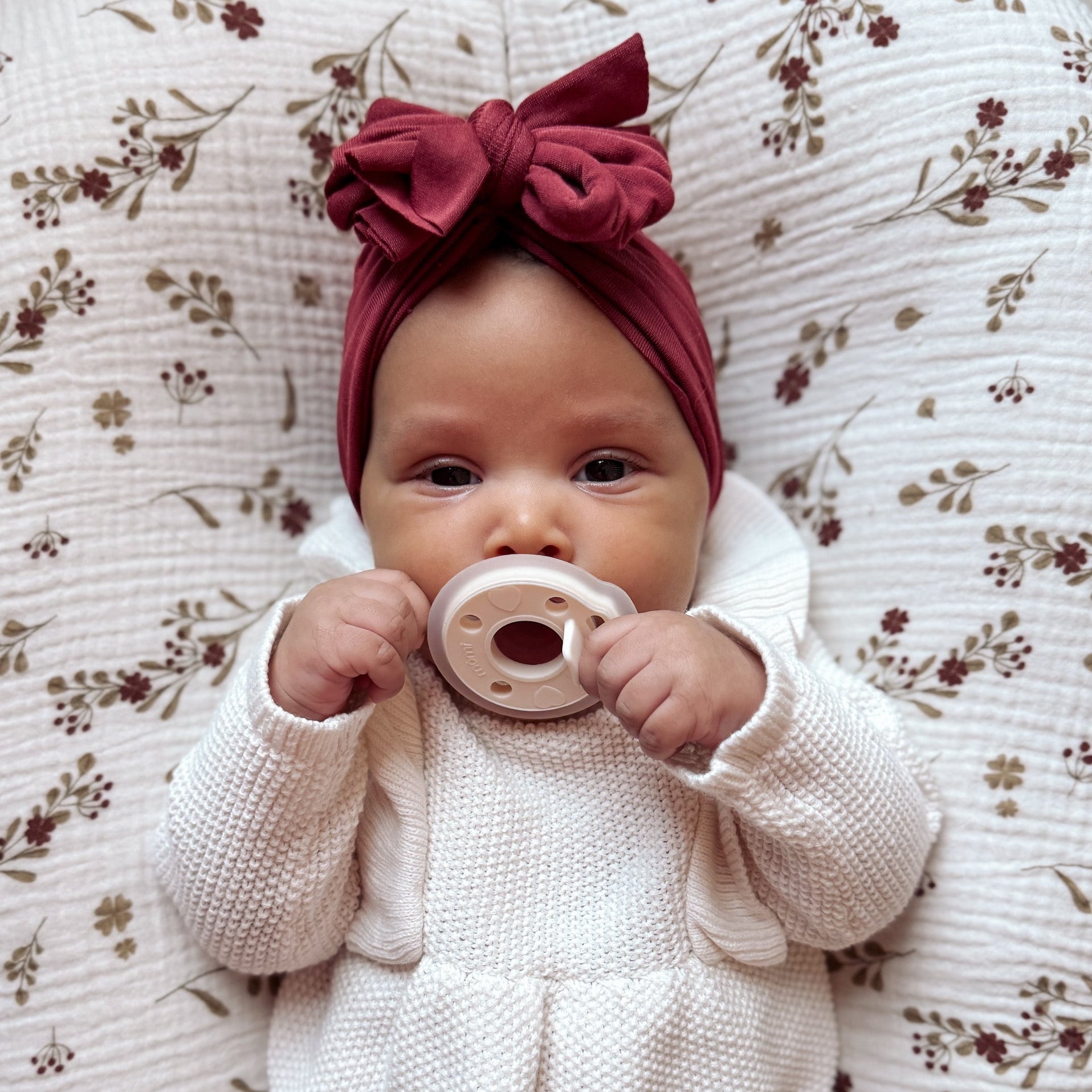 Baby in a white outfit with a red bow headband holding an antique-white "pearl" mōmi pacifier on a floral-patterned cushion