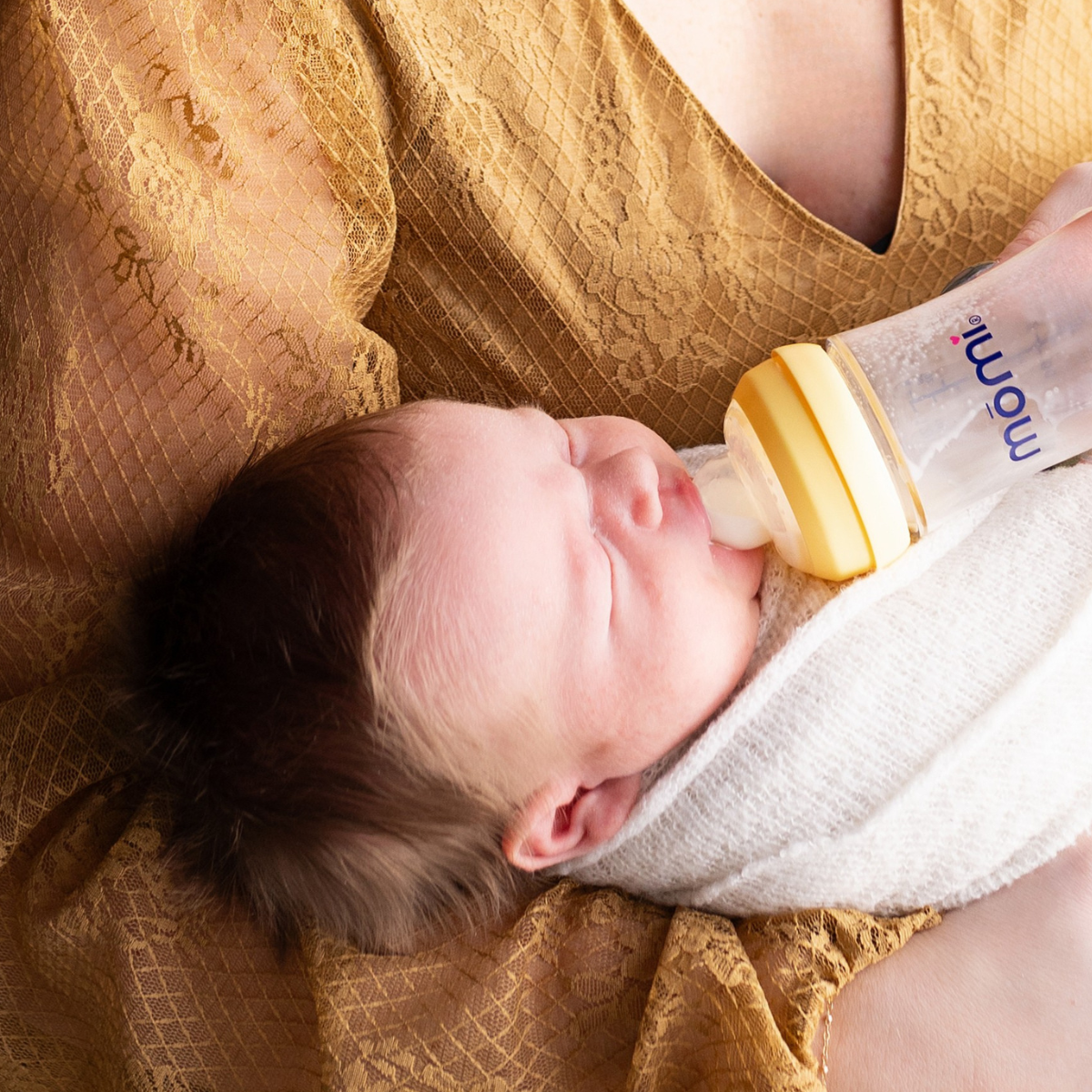 Newborn baby being fed from a mōmi bottle with a sundrop yellow collar in mother's arms